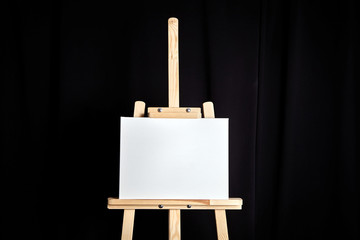 White blank cotton canvas stands on a wooden artistic easel on black curtain background. Horizontal rectangular mockup canvas wrapped on stretcher bar, front view