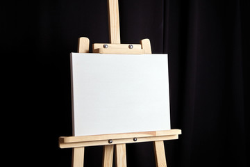 White blank cotton canvas stands on a wooden artistic easel on black curtain background. Horizontal...