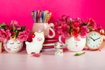Different Notebooks, alarm clock, unicorn mug, llama squishy, Alstroemeria flowers and flamingo mug with colorful gel pens on bright pink background. Idea of Girly Desk table. Back to school concept
