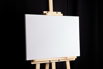 White blank canvas stands on a wooden artistic easel on black curtain background. Horizontal...