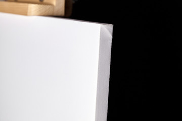 White blank synthetic canvas stands on a wooden artistic easel on black curtain background. Horizontal rectangular mockup canvas wrapped on stretcher bar