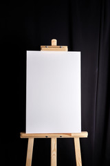 White blank canvas stands on a wooden artistic easel on black curtain background. Vertical...