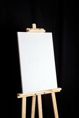 White blank canvas stands on a wooden artistic easel on black curtain background. Vertical...