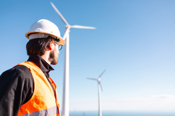 Giovane ingegnere con gilet ad alta visibilità, casco bianco e progetto in mano, sta controllando l'impianto eolico formato da turbine a vento in montagna. Concetto di manutenzione. - obrazy, fototapety, plakaty