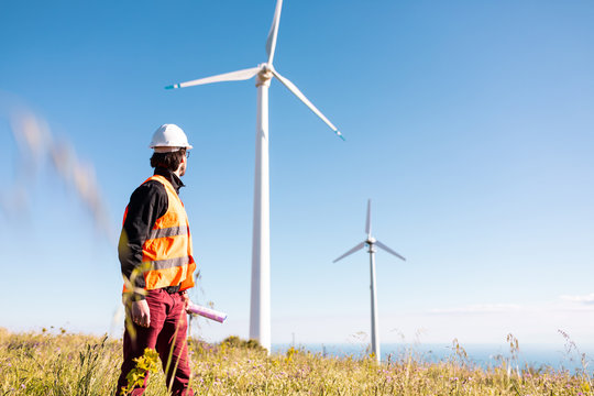 Giovane ingegnere con gilet ad alta visibilit&agrave;, casco bianco e progetto in mano, sta controllando l'impianto eolico formato da turbine a vento in montagna. Concetto di manutenzione.