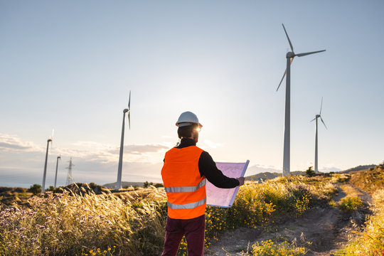 Giovane ingegnere con gilet ad alta visibilit&agrave;, casco bianco e progetto in mano, sta controllando l'impianto eolico formato da turbine a vento in montagna. Concetto di manutenzione.