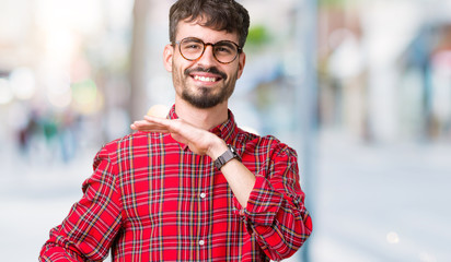 Young handsome man wearing glasses over isolated background gesturing with hands showing big and large size sign, measure symbol. Smiling looking at the camera. Measuring concept.