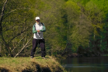 Fisherman throwing the bait into river standing on river bank in sunny day with green forest in background