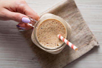 Woman's hand holding a glass jar filled with banana, kiwi, apple smoothie over white wooden surface, top view. Flat lay, from above, overhead.
