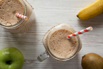 Glass jars filled with banana, kiwi, apple smoothie on a white wooden background. From above, overhead, top view.