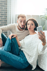selective focus of attractive woman and handsome man with duck faces taking selfie at home