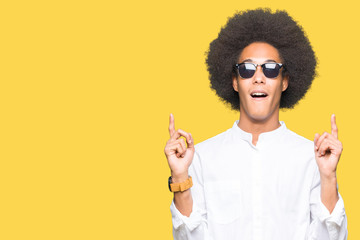Young african american man with afro hair wearing sunglasses amazed and surprised looking up and pointing with fingers and raised arms.
