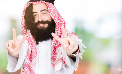 Arabian business man with long hair wearing traditional keffiyeh scarf smiling looking to the camera showing fingers doing victory sign. Number two.