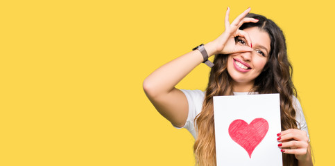 Young adult woman holding card with red heart with happy face smiling doing ok sign with hand on eye looking through fingers