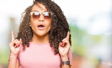 Young beautiful woman with curly hair wearing pink sunglasses amazed and surprised looking up and pointing with fingers and raised arms.