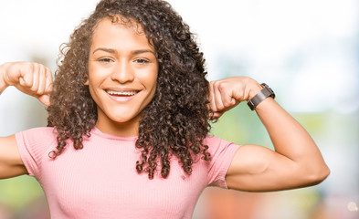 Young beautiful woman with curly hair wearing pink t-shirt showing arms muscles smiling proud. Fitness concept.
