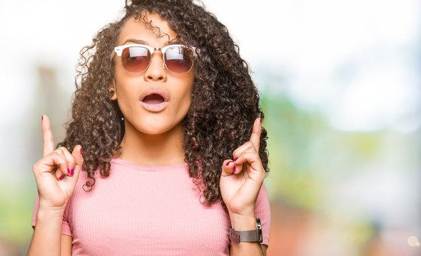 Young beautiful woman with curly hair wearing pink sunglasses amazed and surprised looking up and pointing with fingers and raised arms.