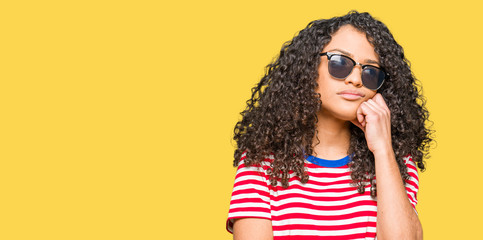 Fototapeta premium Young beautiful woman with curly hair wearing sunglasses with hand on chin thinking about question, pensive expression. Smiling with thoughtful face. Doubt concept.