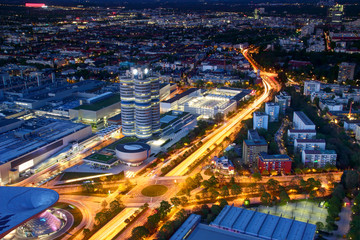 Modern European aerial cityscape in blue hour with broad circle road intersection, commercial, office and industrial buildings in outskirts lit by street and car lights, Munchen Bayern Germany Europe