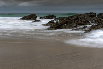 A beach scene with smooth sand and flowing water. Rocks lay across the sand and shoreline