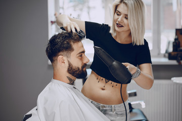 handsome young bearded guy sitting in an armchair in a beauty salon and the girl around him dry his hair