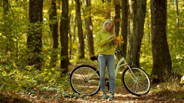 Autumn woman with retro bike with flowers in basket in autumnal park. Autumn happy girl and joy. Carefree woman. Outdoor atmospheric fashion photo of young beautiful lady in autumn landscape.