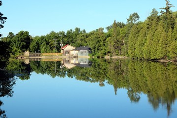 Obraz premium Perfect reflection of wood buildings and treeline on the bright blue water at the top of the dam in Scone near Chesley, Ontario, Canada on a calm, clear, bright sunny morning.
