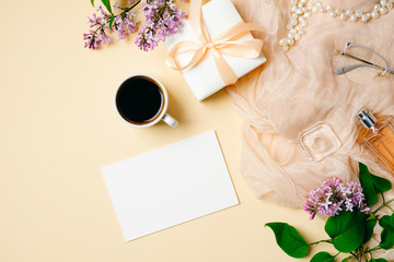 Flat lay, top view office table desk. Women workspace with paper card, coffee cup, lilac flowers bouquet, gift box, perfume bottle on beige background. Feminine desktop. Wedding invitation concept.