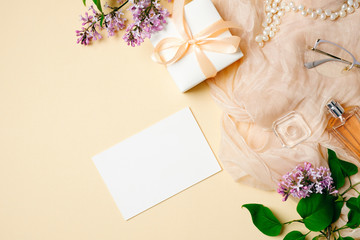 Top view, flat lay womens desk. Lilac flowers, silk scarf, glasses, perfume bottle, gift box and blank greeting card on beige feminine background. Elegant and glamour female stuff. Wedding invitation.