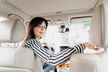 Attractive young brunette woman sitting behind a wheel of a car sitting and smiling looking at a back seat where her children sit