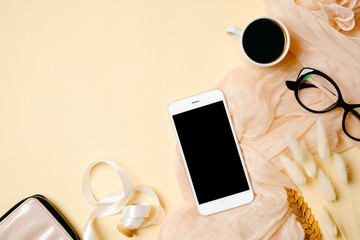Woman home office desk with mobile phone, coffee cup, glasses, silk scarf and accessories on beige background. Flat lay beauty blogger workspace. Top view feminine background.