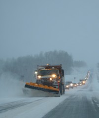 Snowplow clearing away drifting snow on Ontario highway in January 2019 at dusk with long line of vehicles following behind