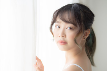 Portrait of beautiful young asian woman standing at the window and smile while wake up with sunlight at morning, girl happy with fresh and cheerful, lifestyle and relax for wellness concept.