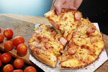 Hot homemade pizza slice with the man hand and melting cheese, sausage on top and small tomato on wooden board on black table background. Foods concept.
