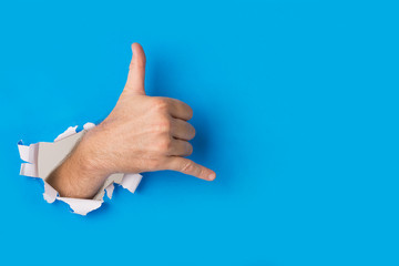 Male hand tearing through blue paper background creating a shaka hand gesture.