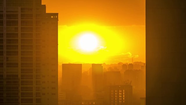 Big orange sunset twilight time lapse from behind clouds in urban landscape with buildings silhouettes and strong light using telephoto lens feeling very hot during summer in Sao Paulo City downtown