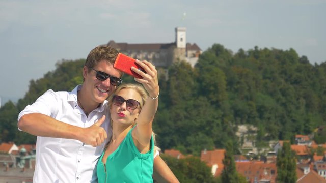 HANDHELD, CLOSE UP: Carefree girl kisses her handsome boyfriend on the cheek while filming a travelling v-log. Cheerful couple taking selfies from a rooftop in picturesque Ljubljana city center.