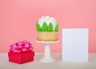 Large cup cake with giant frosting rose sitting on white pedestal on wood table with bright present and blank card, copy space