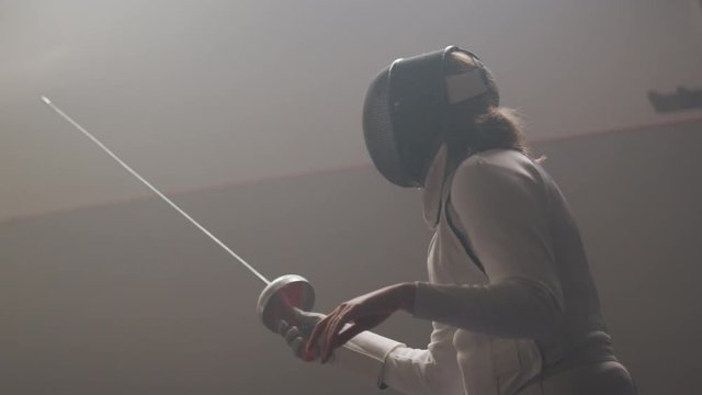 A female fencer raises her foil in a dark and dusty fencing gym in slow motion