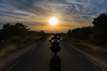A heavy motorcycle with red lights driving a narrow and completely straight road into the sunset on the horizon with mountains.