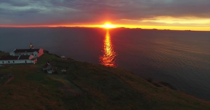 Aerial view of Bruce lighthouse on a rocky cliff of grey basalt stone. Stunning sunrise as bright as fire and dramatic cloudy sky above the sea. Far East of Russia