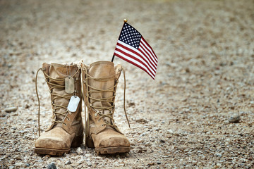 Old military combat boots with dog tags and a small American flag. Rocky gravel background with copy space. Memorial Day, Veterans day, sacrifice concept. - obrazy, fototapety, plakaty