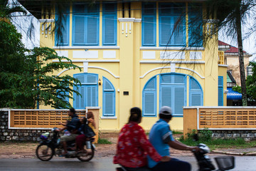 Khmer people riding motorcycle past french colonial building. 