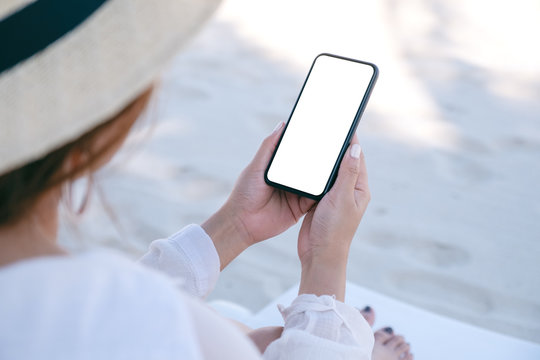 Mockup image of a woman holding and using white mobile phone with blank desktop screen while sitting on beach chair on the beach