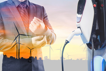 double exposure businessmen and silhouette of wind turbine at sunset with electric car battery charger station. Green energy for new innovative technology EV Electrical vehicle concept.