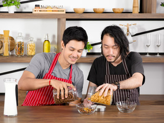 Asian young men wear apron are pouring cereal into a glass bowl together on wooden table in modern dining room apartment on weekend.
