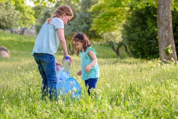 Fototapeta premium Cute little girls cleaning up plastic litter on grass. Children Volunteers cleaning up litter and putting plastic bottle into recycling bag. World Environment Day.