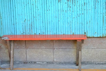 An empty old orange woiden bench on the ground floor with a brick wall and blue zinc wall