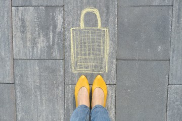 Shopping bag picture written on gray sidewalk in crayons with women legs in yellow shoes