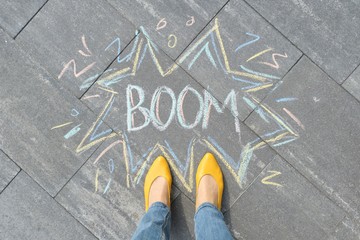 Boom written on gray sidewalk with women legs in yellow shoes